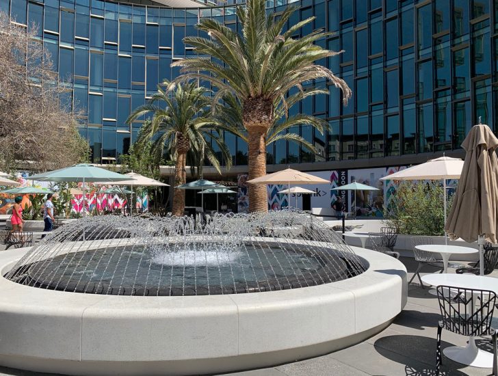 Wide bench seating and shaded dining tables around Santana Row Plaza entrance fountain